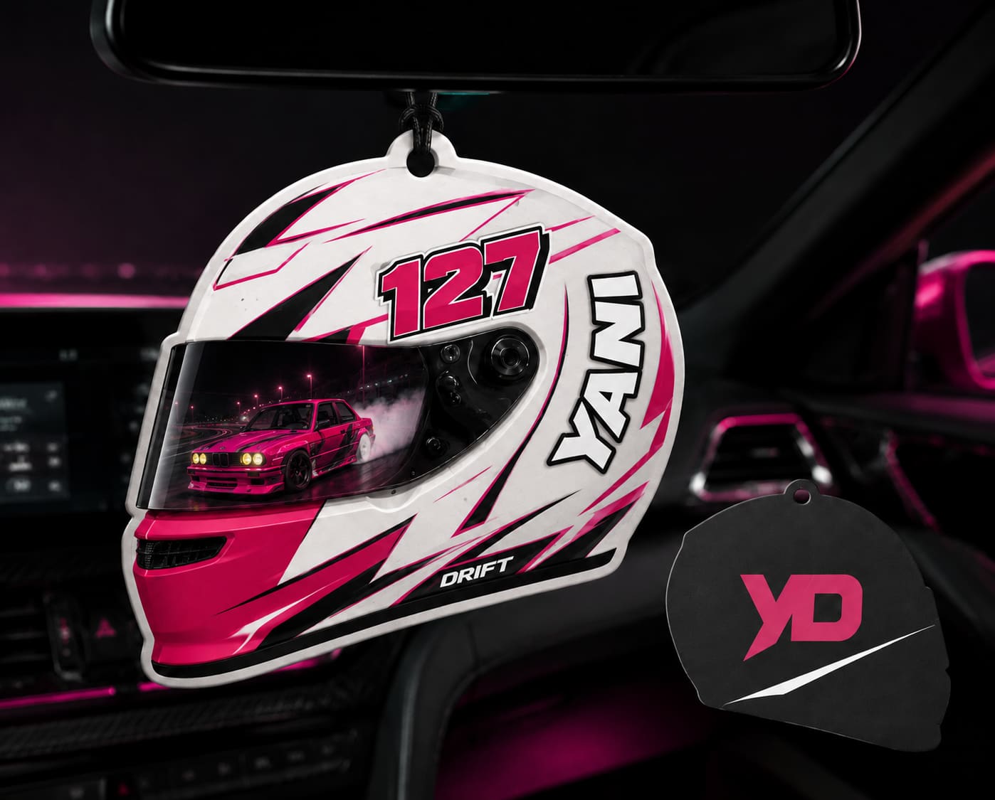 Ambientador con forma del casco de Yanibel Díaz en blanco y rosa, con el 127 y el nombre YANI, un BMW E30 derrapando reflejado en la visera, y el reverso negro con el monograma YD